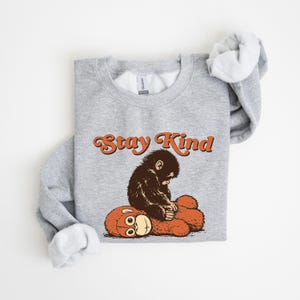 May include: A gray sweatshirt with the words "Stay Kind" in orange script. The graphic features a brown monkey sitting on a red teddy bear. The sweatshirt has a crew neck and long sleeves.