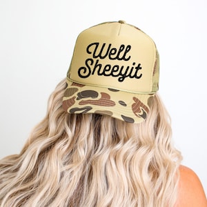 May include: A tan trucker hat with a camouflage pattern on the brim and the text "Well Sheeeyit" in black script. The hat has a mesh back and a curved brim. The hat is worn by a person with long, wavy blonde hair.