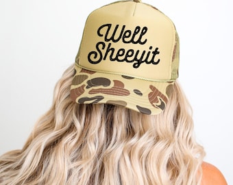 May include: A tan trucker hat with a camouflage pattern on the brim and the text "Well Sheeeyit" in black script. The hat has a mesh back and a curved brim. The hat is worn by a person with long, wavy blonde hair.