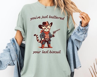 May include: A pale green t-shirt with a cartoon cowboy cat holding a gun. The cat wears a hat, red neckerchief, and boots. The text on the shirt reads "you've just buttered your last biscuit."