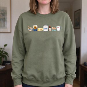 May include: An olive green crewneck sweatshirt featuring a graphic print of baking ingredients in a row, including a mixing bowl, a bag of Gold Medal flour, a stick of butter, a jar labeled SUGAR, vanilla extract, eggs, and a measuring cup.