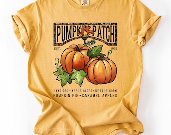 Op de afbeelding: Mosterdgeel T-shirt met een afbeelding van twee oranje pompoenen en groene bladeren, en de tekst "PUMPKIN PATCH". Het shirt vermeldt ook "HAYRIDES, APPLE CIDER, KETTLE CORN, PUMPKIN PIE, CARAMEL APPLES."