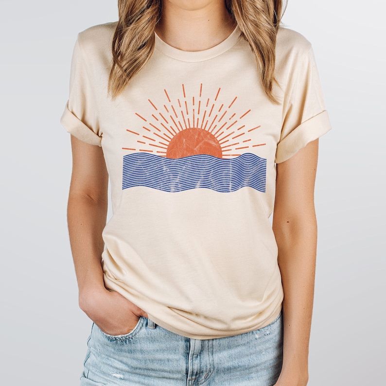 May include: A beige t-shirt with a graphic of a red sun setting over blue waves. The sun has a textured design and the waves are made up of thin blue lines.