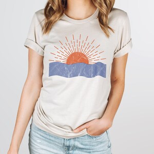 May include: A beige t-shirt with a graphic of a red sun setting over blue waves. The sun has a red outline and orange fill. The waves are blue with a white outline.