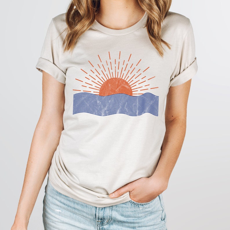 May include: A beige t-shirt with a graphic of a red sun setting over blue waves. The sun has a red outline and orange fill. The waves are blue with a white outline.