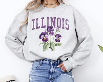 May include: Gray sweatshirt with the word "ILLINOIS" printed in purple on the front. The sweatshirt features a purple and green floral design with three purple pansies.