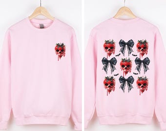 May include: A light pink sweatshirt featuring a strawberry skull design. The front has a single strawberry skull, while the back showcases multiple strawberry skulls with black bows and bat accents. The design is a unique blend of cute and spooky.