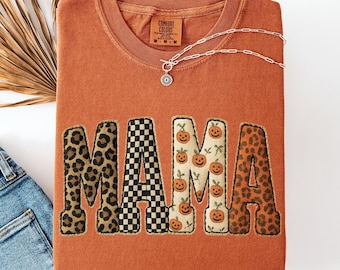 Pode incluir: Uma camiseta laranja Comfort Colors com a palavra "MAMA" em um design de patchwork. As letras apresentam estampas de leopardo, xadrez preto e branco e padrões de abóbora. Um colar de prata com um pingente é drapeado sobre a camisa.