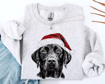 Può includere: Una felpa grigio chiaro con un'illustrazione in bianco e nero di un Labrador Retriever con un cappello da Babbo Natale rosso e bianco. La felpa ha maniche lunghe e girocollo, perfetta per le festività.