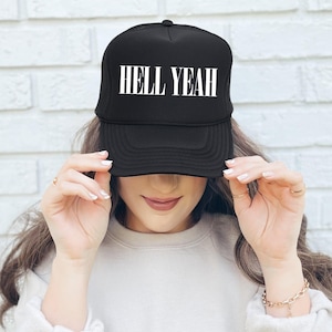 Könnte beinhalten: Schwarze Trucker-Mütze mit dem weißen Text "HELL YEAH" auf der Vorderseite. Die Mütze hat eine Netzrückseite und einen gebogenen Schirm. Die Mütze wird von einer Person gehalten.