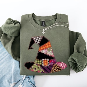 May include: An olive green sweatshirt featuring a patchwork witch hat design in various patterns and colours. A silver necklace with a circular pendant rests on the shirt. The sweatshirt is folded on top of blue jeans.
