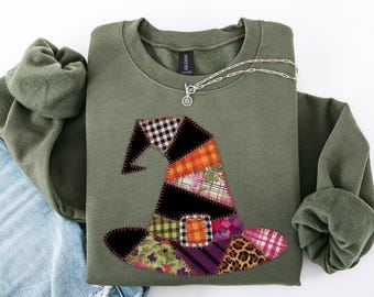 May include: An olive green sweatshirt with a patchwork witch hat design in various patterns and colors. A silver necklace with a circular pendant rests on the shirt. The sweatshirt is folded on top of a pair of blue jeans.