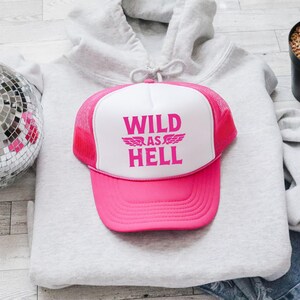 Op de afbeelding: Een roze en witte truckerpet met de tekst "WILD AS HELL" in roze. De pet heeft een roze klep en een mesh achterkant. De pet ligt op een grijze hoodie.