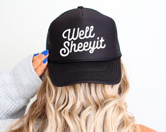 May include: Black trucker hat with the text "Well Sheeyit" in white script. The hat has a mesh back and a curved brim. The hat is being worn by a person with blonde hair.