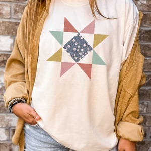 Op de afbeelding: Crèmekleurig T-shirt met een kleurrijk sterrenpatroon. De ster heeft driehoeken in roze, groen en geel, met een centraal vierkant van blauwe stof met een bloemenpatroon. Het shirt heeft een relaxte pasvorm.
