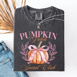 May include: Dark gray t-shirt with the words "PUMPKIN Patch Social Club" in pink and orange. The shirt features a watercolor-style pumpkin with a pink bow and a leafy design. A silver necklace with a pendant is draped on the shirt.