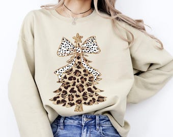 May include: A beige sweatshirt with a Christmas tree design. The tree is decorated with a leopard print pattern and a bow with black polka dots. A gold glitter star sits atop the tree. The sweatshirt is paired with blue jeans.