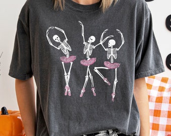 Può includere: Una maglietta grigia scura con una grafica di tre scheletri vestiti da ballerine. Gli scheletri indossano tutù rosa e sono in varie pose di danza.