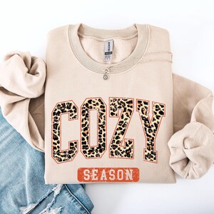 May include: A beige sweatshirt with the words "COZY SEASON" in a leopard print design. The sweatshirt is folded and has a silver necklace with a small pendant. The sweatshirt is on a white surface with a pair of blue jeans.