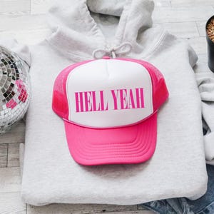Könnte beinhalten: Eine pinkfarbene und weiße Trucker-Cap mit dem Text "HELL YEAH" in kräftigen pinkfarbenen Buchstaben. Die Cap liegt auf einem grauen Kapuzenpullover. Die Cap hat ein Mesh-Rückenteil und einen gebogenen Schirm. Das Thema ist lässig und lustig.