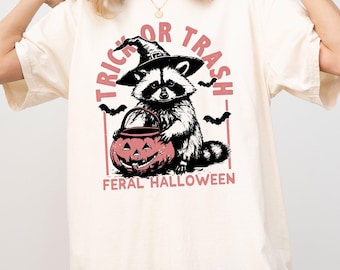 Op de afbeelding: Crèmekleurig T-shirt met een wasbeer met een heksenhoed en een roze pompoen. De tekst "TRICK OR TRASH" en "FERAL HALLOWEEN" is roze gedrukt. Zwarte vleermuizen zijn ook inbegrepen.