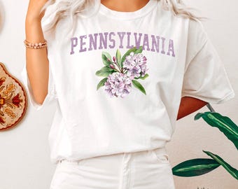 May include: White t-shirt featuring the word "PENNSYLVANIA" in purple lettering, above a floral design of purple flowers and green leaves. A casual style t-shirt.