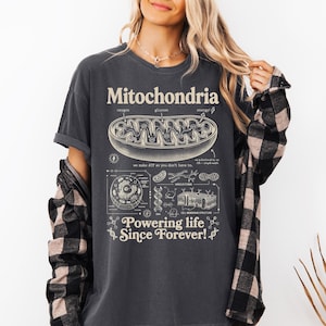 May include: Dark gray t-shirt featuring a detailed diagram of a mitochondria, with labels for oxygen, glucose, and energy. The text reads "Mitochondria" and "Powering life Since Forever!" A science-themed graphic tee.