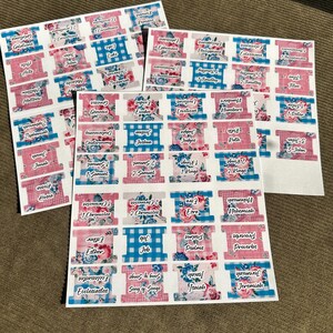 May include: Sheets of Bible tabs with various book names printed on them. The tabs feature floral, gingham, and solid color designs in pink, blue, and white. The text is in a handwritten style font.