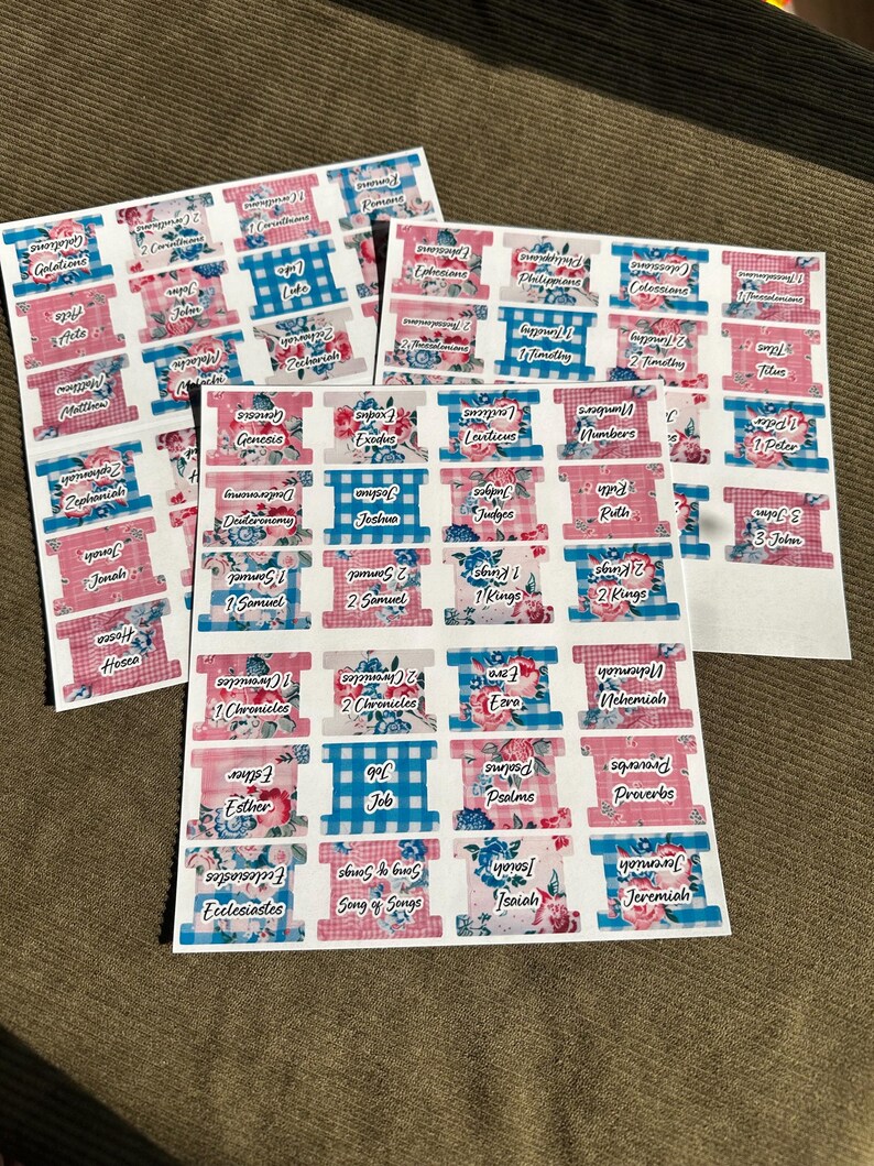 May include: Sheets of Bible tabs with various book names printed on them. The tabs feature floral, gingham, and solid color designs in pink, blue, and white. The text is in a handwritten style font.