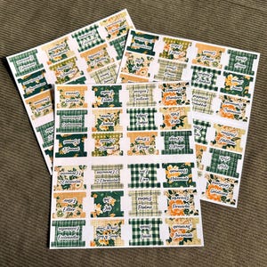 May include: Sheets of stickers with Bible book names. Each sticker features a decorative design with floral and plaid patterns in green, gold, and white. The stickers are arranged on a neutral background.
