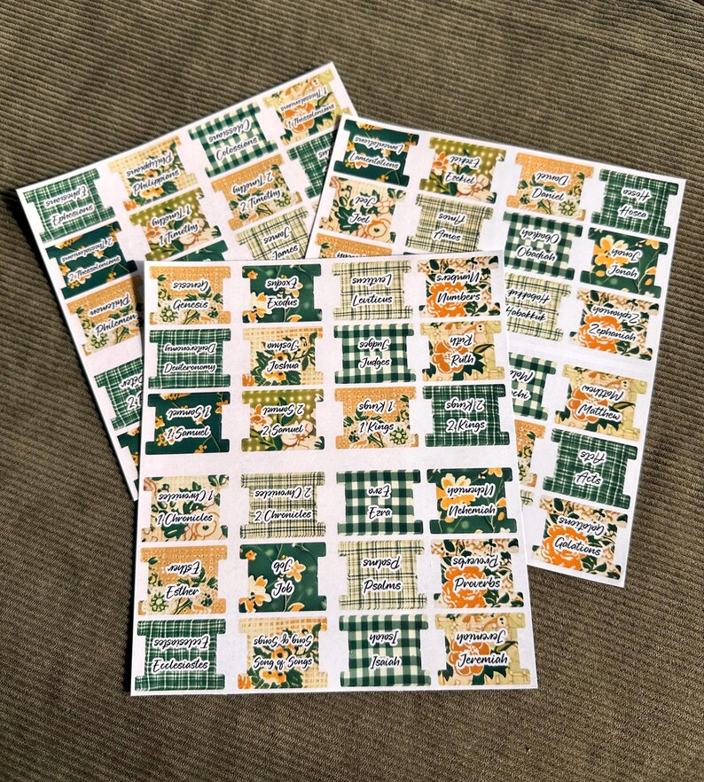 May include: Sheets of stickers with Bible book names. Each sticker features a decorative design with floral and plaid patterns in green, gold, and white. The stickers are arranged on a neutral background.