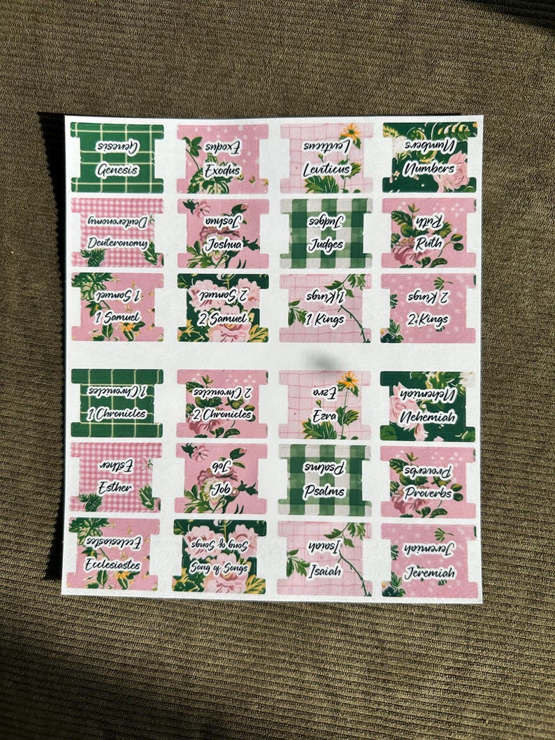 May include: A sheet of decorative Bible book labels. The labels feature a mix of floral and checkered patterns in pink and green. Each label displays a book of the Bible in a handwritten font.