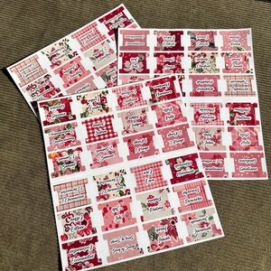May include: Sheets of Bible tabs with various floral and plaid patterns in shades of pink and red. Each tab is labeled with a book of the Bible in a white font. The tabs are arranged on a brown surface.