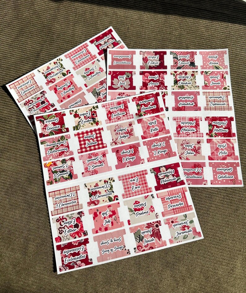 May include: Sheets of Bible tabs with various floral and plaid patterns in shades of pink and red. Each tab is labeled with a book of the Bible in a white font. The tabs are arranged on a brown surface.