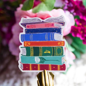 May include: A sticker featuring a stack of colorful books. The books are in various colors, including pink, blue, red, green, and teal. The top book is open, and the sticker is set against a floral background.