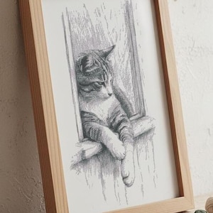 May include: A framed monochrome print of a cat in a window. The cat is depicted in shades of grey and white, resting on a window ledge. The frame is made of light-coloured wood.