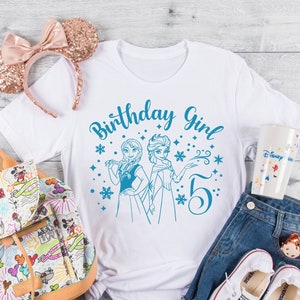 Könnte beinhalten: Ein weißes T-Shirt mit einem blauen Design, auf dem "Birthday Girl" steht, sowie ein Bild von Elsa und Anna aus Frozen und die Zahl 5. Das Design ist von Schneeflocken und Sternen umgeben.
