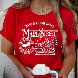 May include: Red t-shirt with white text that reads "Baked Fresh Daily Main Street Cookie Co. Serving Orlando Since 1971". The shirt features a white Mickey Mouse head with a cookie in his hand.