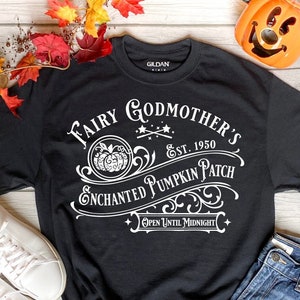 Peut inclure: T-shirt noir avec un texte blanc qui dit "Fairy Godmother's Enchanted Pumpkin Patch Est. 1950 Open Until Midnight". Le texte est dans une police de caractères vintage avec un graphique de citrouille.