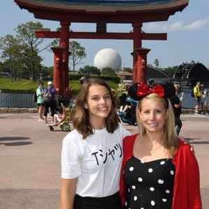 May include: Two people, a young woman wearing a white t-shirt with the Japanese text "Tシャツ" and a black skirt, and an older woman wearing a red cardigan over a black and white polka dot dress, stand in front of a red Japanese torii gate. The gate is decorated with a blue roof and a white sphere is visible in the background.
