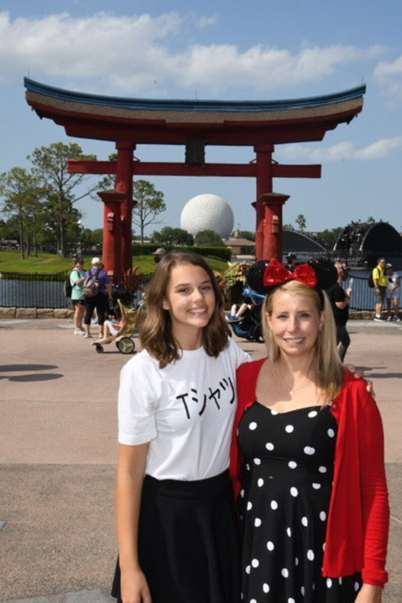 May include: Two people, a young woman wearing a white t-shirt with the Japanese text "Tシャツ" and a black skirt, and an older woman wearing a red cardigan over a black and white polka dot dress, stand in front of a red Japanese torii gate. The gate is decorated with a blue roof and a white sphere is visible in the background.