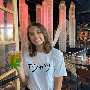 May include: A woman wearing a white t-shirt with the Japanese text "T-シャツ" printed on it. She is holding a green and yellow layered drink with a purple straw. The background is a restaurant with a modern design.