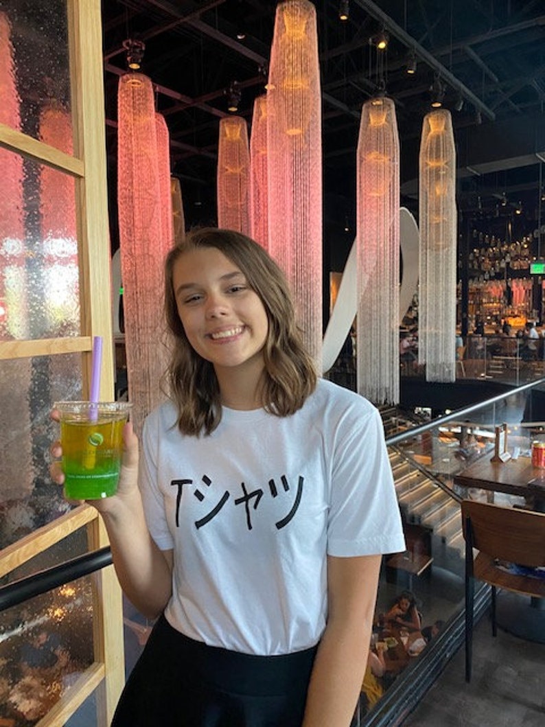 May include: A woman wearing a white t-shirt with the Japanese text "T-シャツ" printed on it. She is holding a green and yellow layered drink with a purple straw. The background is a restaurant with a modern design.