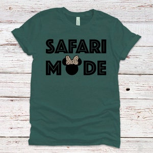 K&ouml;nnte beinhalten: Ein dunkelgr&uuml;nes T-Shirt mit dem Text "SAFARI MODE" in schwarzen umrandeten Buchstaben. Eine schwarze Silhouette eines Mickey Mouse Kopfes mit einer Schleife im Leopardenmuster ist zwischen den W&ouml;rtern zentriert.