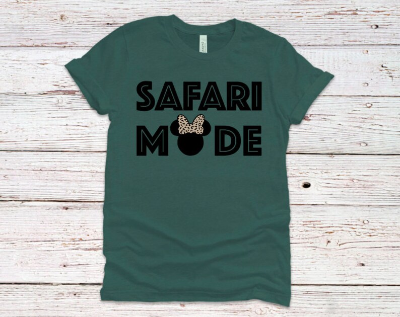 K&ouml;nnte beinhalten: Ein dunkelgr&uuml;nes T-Shirt mit dem Text "SAFARI MODE" in schwarzen umrandeten Buchstaben. Eine schwarze Silhouette eines Mickey Mouse Kopfes mit einer Schleife im Leopardenmuster ist zwischen den W&ouml;rtern zentriert.
