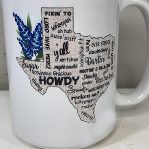 May include: White ceramic mug featuring a Texas-shaped design with the word "Howdy" and other Texan slang terms. The mug also includes a bluebonnet floral illustration, a state flower of Texas.