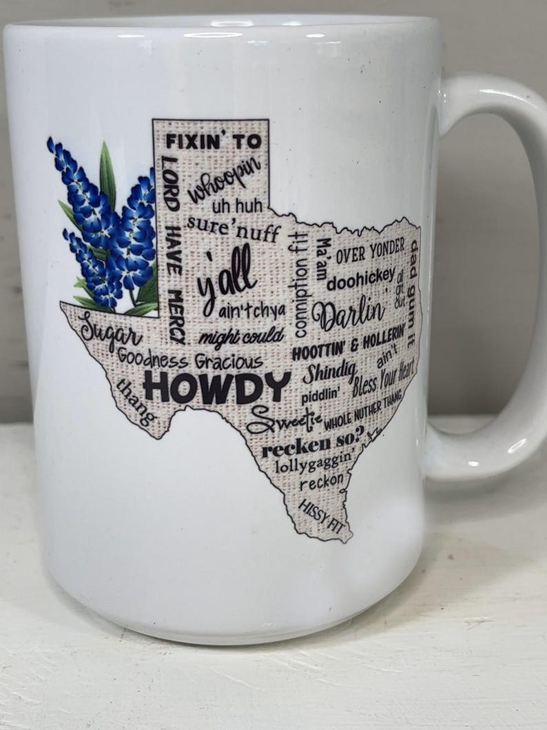 May include: White ceramic mug featuring a Texas-shaped design with the word "Howdy" and other Texan slang terms. The mug also includes a bluebonnet floral illustration, a state flower of Texas.