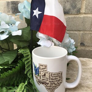 May include: A white ceramic mug with a Texas-shaped graphic featuring the words "Howdy" and "Y'all", and other Texas slang. A small Texas flag is placed behind the mug. The mug has a large handle and is surrounded by greenery and blue flowers.