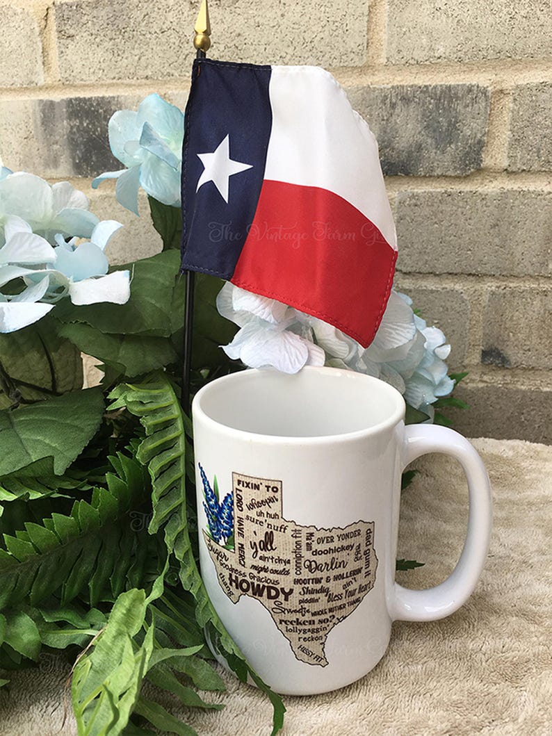 May include: A white ceramic mug with a Texas-shaped graphic featuring the words "Howdy" and "Y'all", and other Texas slang. A small Texas flag is placed behind the mug. The mug has a large handle and is surrounded by greenery and blue flowers.