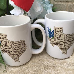 May include: Two white ceramic mugs with a Texas-shaped design. The design includes the words "Howdy", "Y'all", and "Darlin'", along with a bluebonnet flower illustration. The mugs have a curved handle.